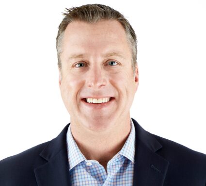 A man wearing a navy blazer and a light blue checkered shirt smiles at the camera against a white background.
