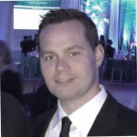 A man in a suit and tie stands indoors at a formal event with people and decorated tables in the background.
