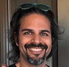 A man with long dark hair, a beard, and mustache smiles at the camera. He is wearing sunglasses on his head and standing indoors.