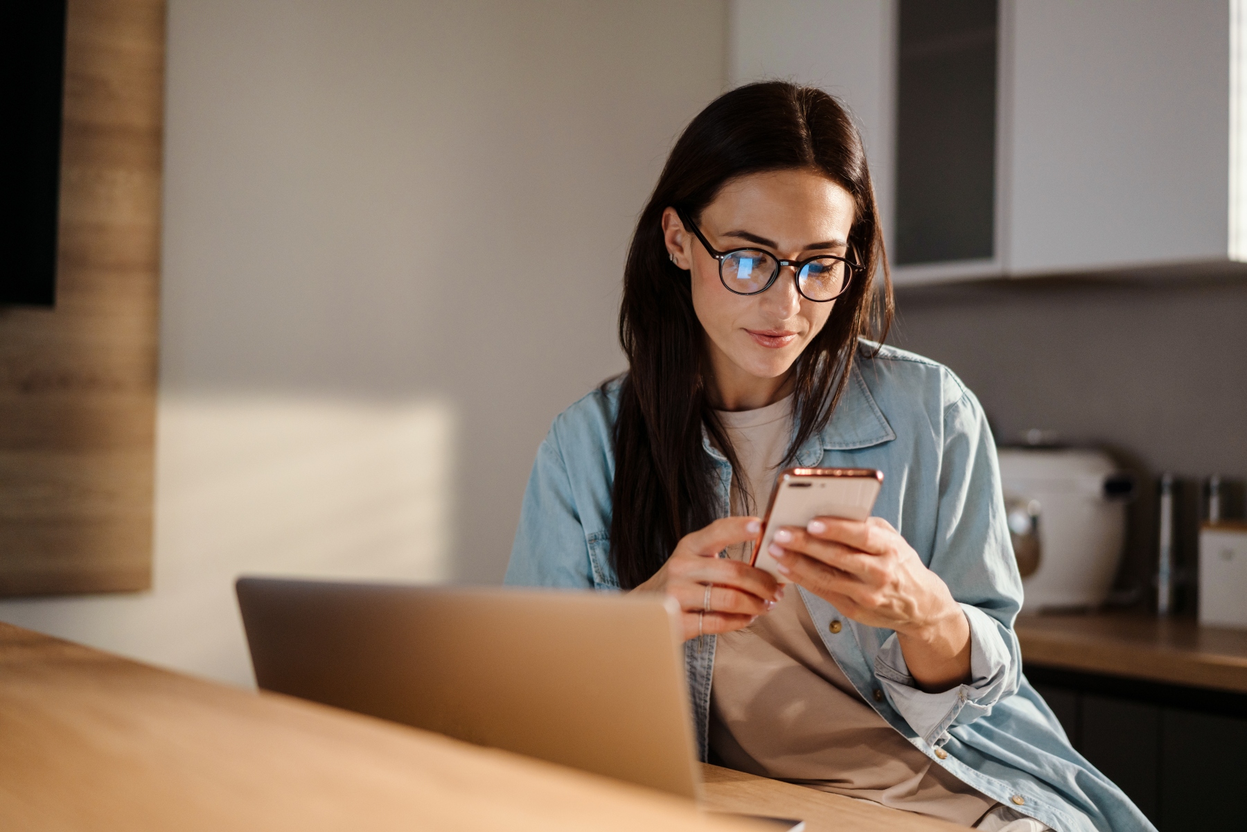 A woman wearing glasses sits at a table with a laptop, looking at her smartphone in a modern kitchen.