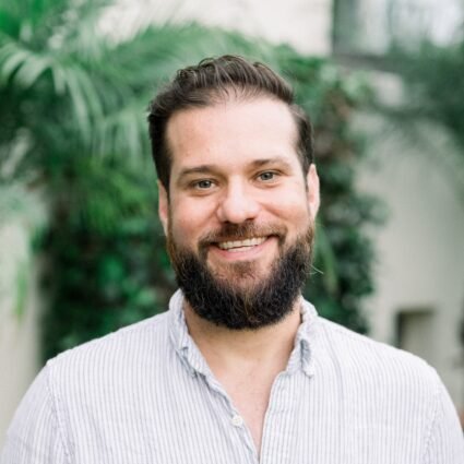 A man with dark hair and a beard, wearing a light striped shirt, smiles outdoors with green foliage in the background.