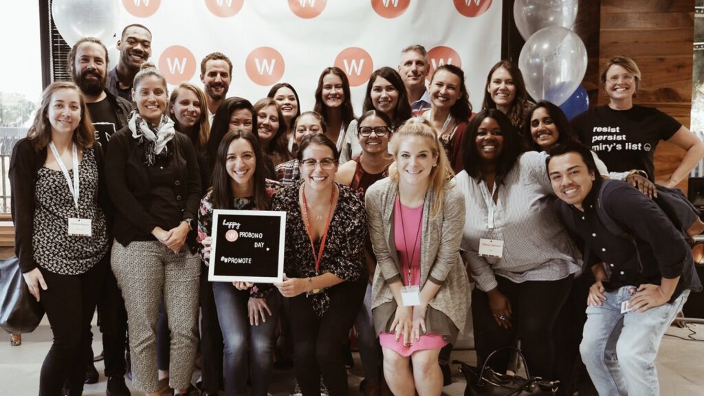 A group of people pose together indoors, some wearing name badges. Two people in front hold a small sign that says "Happy Friends Day #WICOTALENT.