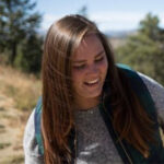 A young woman with long brown hair smiles while standing outdoors on a sunny day, with trees and open land in the background.