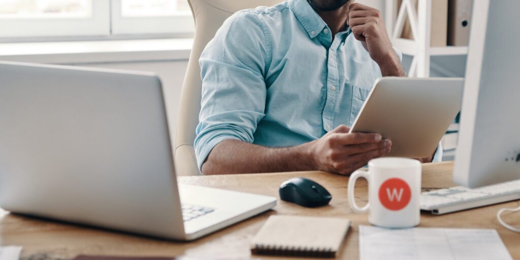 Man sitting at a desk holding a tablet, with a laptop, notebook, mug, and computer monitor in front of him.