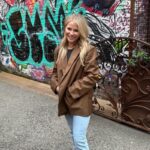 A woman with long blonde hair wearing a brown blazer and light jeans stands and smiles in front of a graffiti-covered wall and metal gate, embodying creative style perfect for an ecommerce marketing campaign.