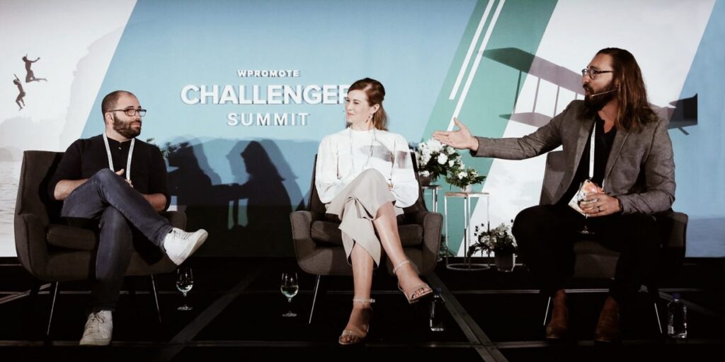 Three people sit on stage having a panel discussion at the Wpromote Challenger Summit, with microphones and event signage in the background.