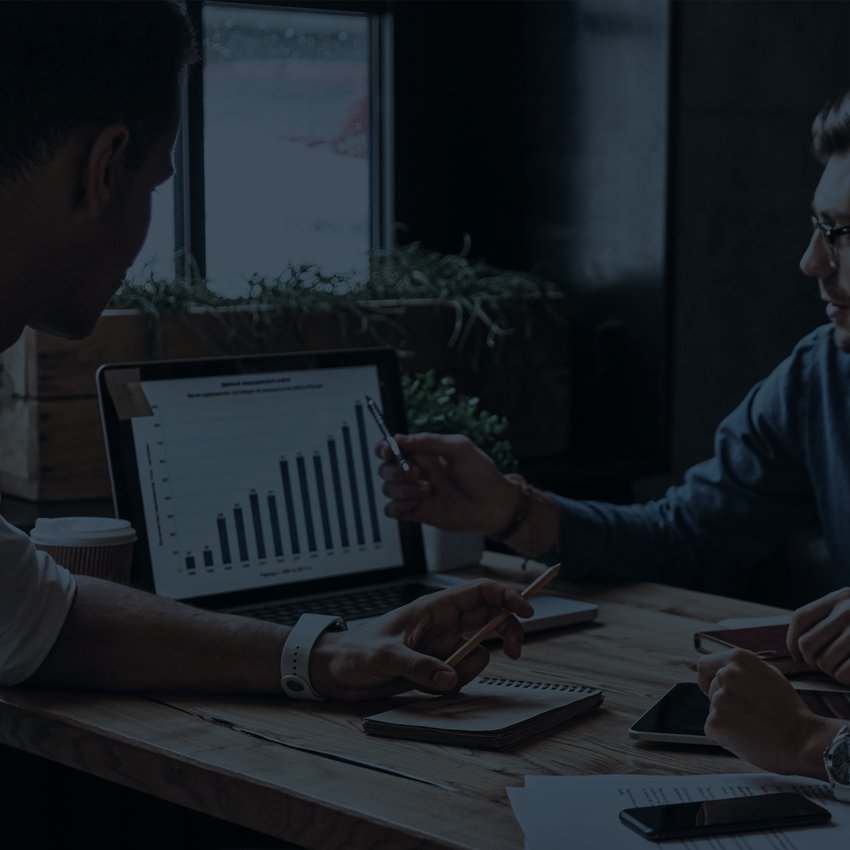 Two people sit at a table discussing a bar graph displayed on a laptop screen, with notebooks, a phone, and papers on the table.