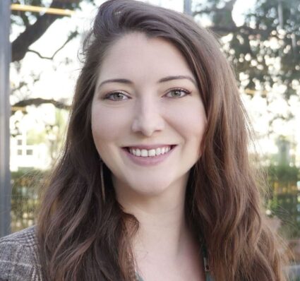 A woman with long brown hair smiling outdoors, with trees and a building blurred in the background.