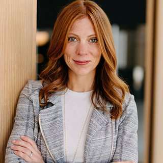 A woman with long red hair, wearing a gray jacket and white shirt, stands indoors with her arms crossed, leaning against a wooden wall.