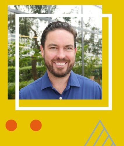 A man with short dark hair and a beard, wearing a blue shirt, smiles while standing in front of greenery and large windows. The image has a yellow graphic border.