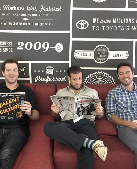 Three men sit on a red couch against a wall with business-themed graphics; two are reading magazines and one is smiling at the camera.