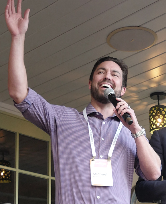 A man wearing a name badge speaks into a microphone and raises his left arm while standing indoors under a paneled ceiling.