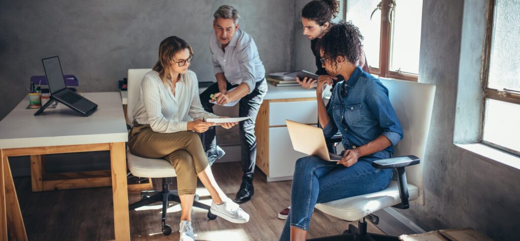 Four people in an office setting sit and stand around desks, discussing documents and using a laptop under natural light from a window as they collaborate on marketing strategy services.