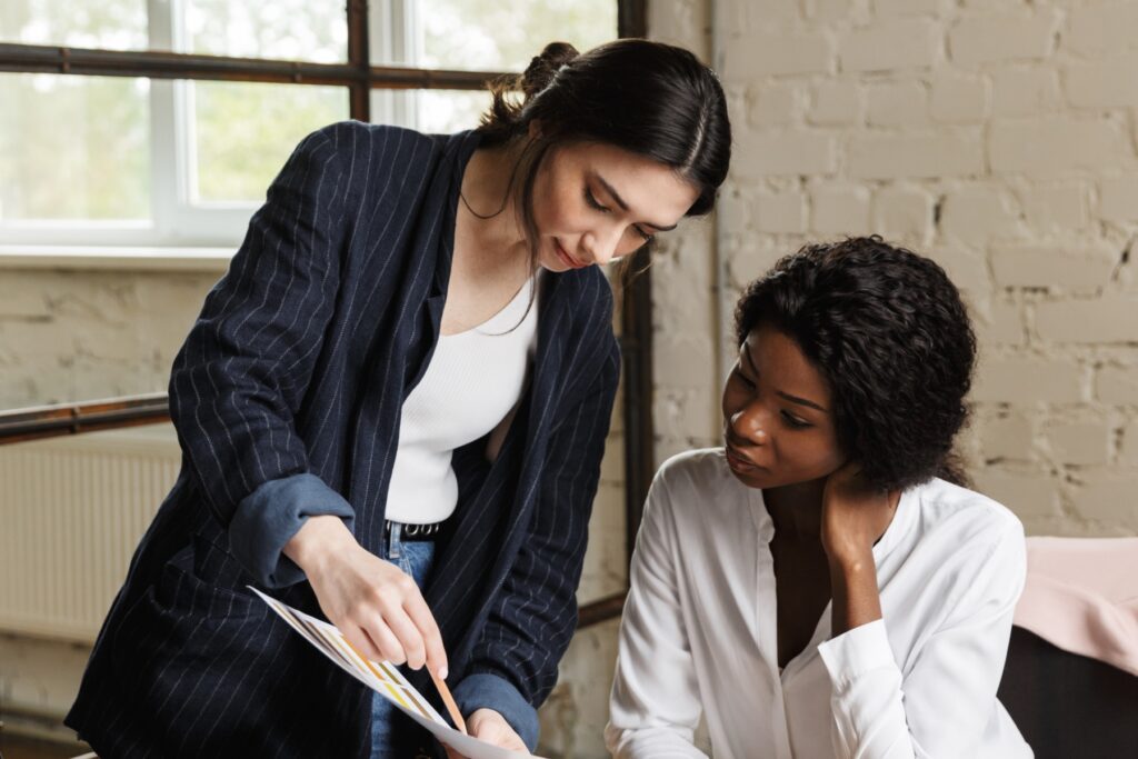 Two women in business attire review a document together in a modern office with a white brick wall and large window.