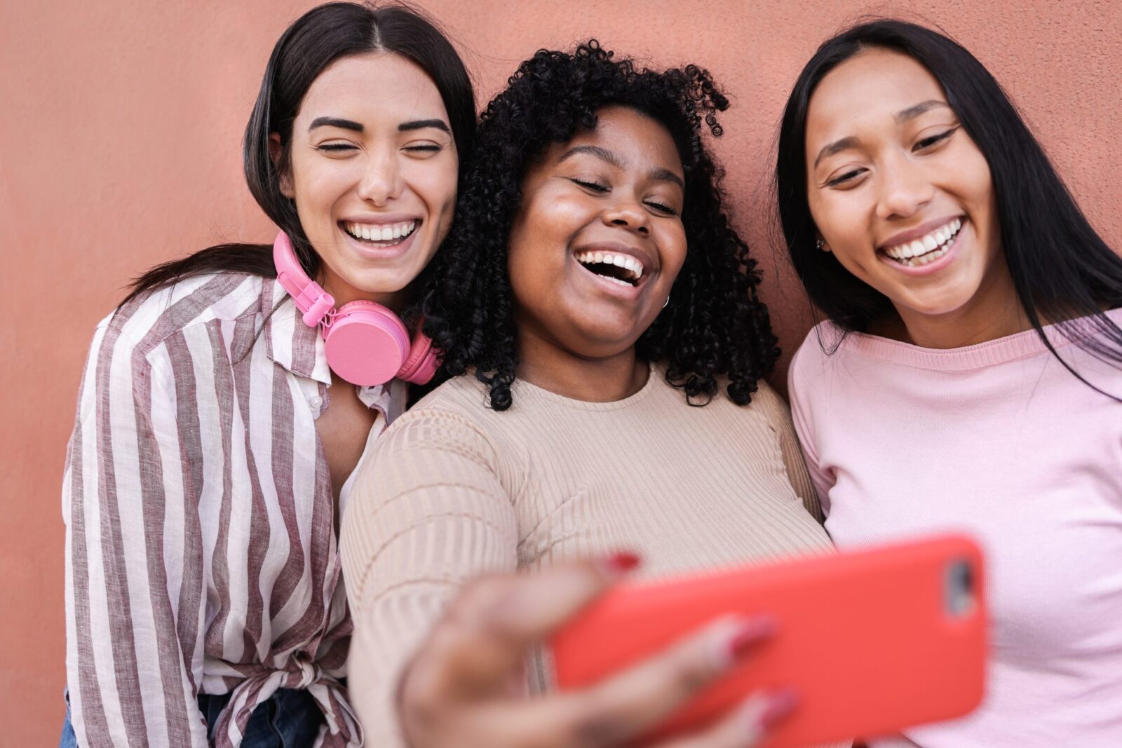 Three young women stand close together, smiling and taking a selfie against a pink wall—capturing the fun side of friendship and great marketing strategy services. One wears headphones around her neck.