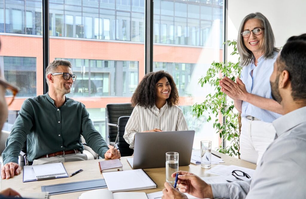 Four people sit at a conference table with laptops and notebooks while a woman stands and discusses digital marketing services, all appearing engaged in a modern office setting with large windows.