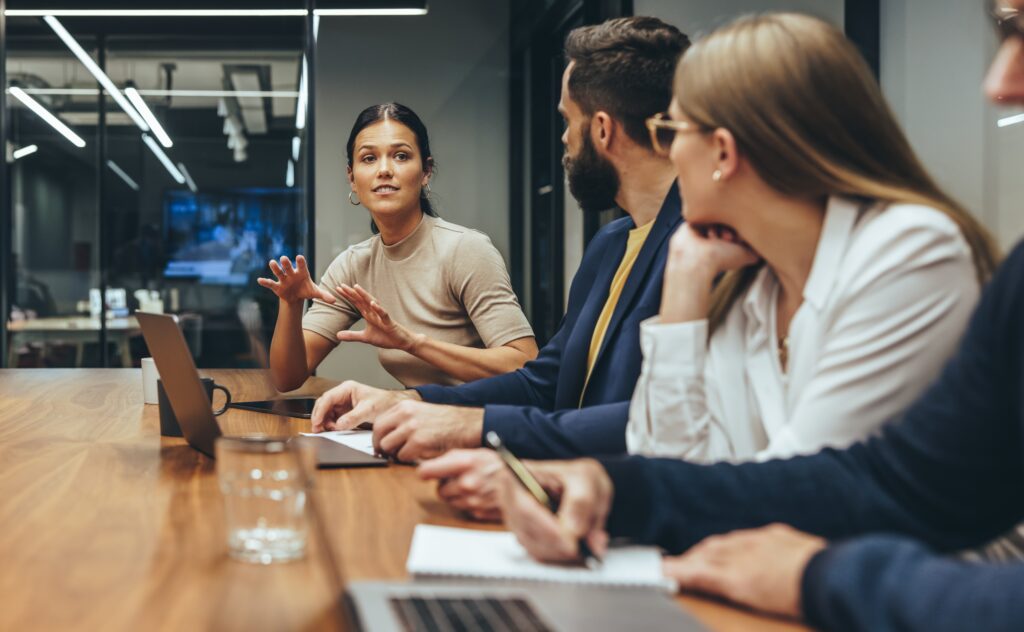 Four people sit at a conference table in an office, discussing digital marketing services as one woman speaks while the others listen and take notes.