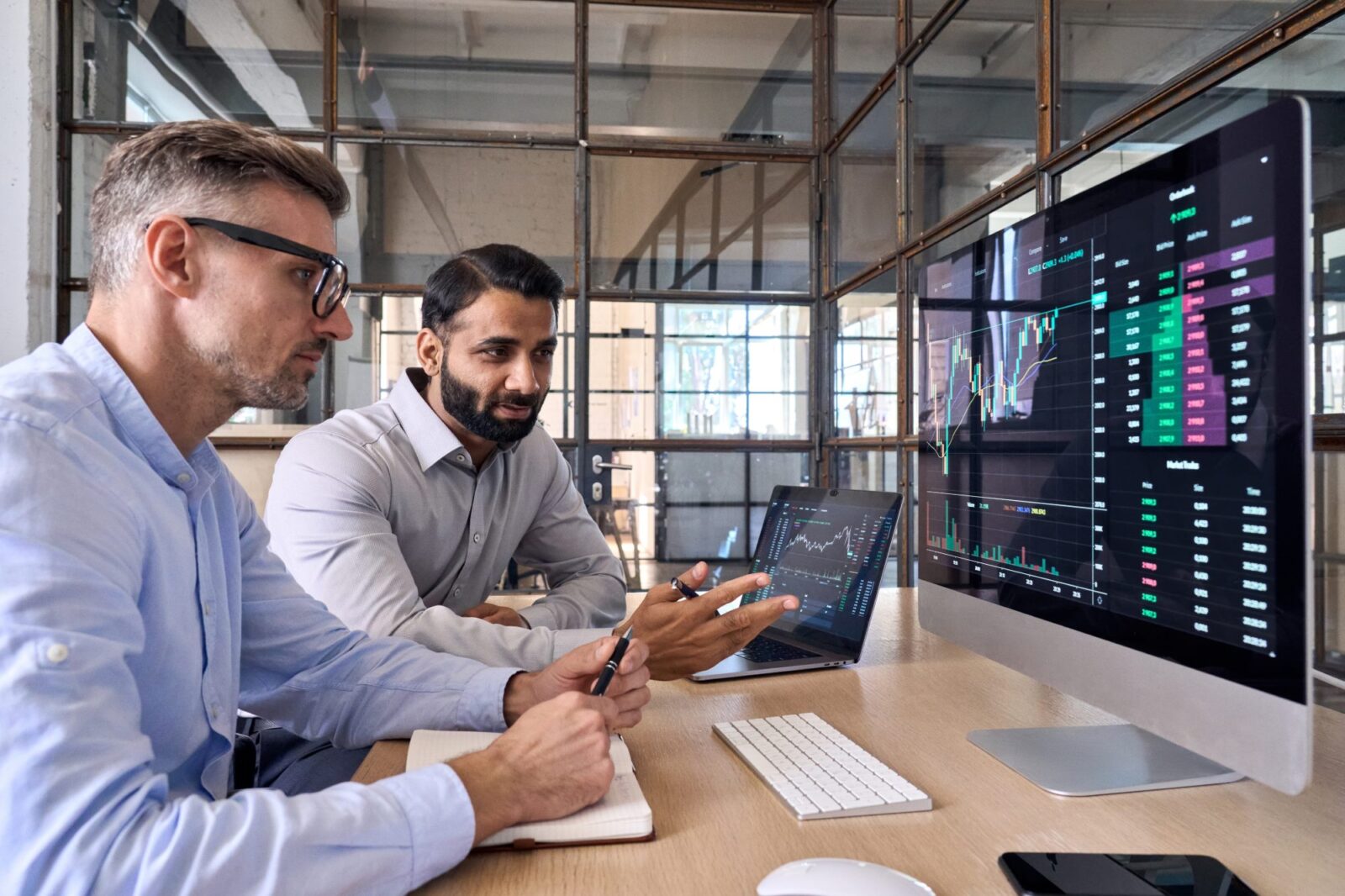 Two men sit at a desk analyzing financial data and stock charts displayed on a large computer monitor in a modern office setting.