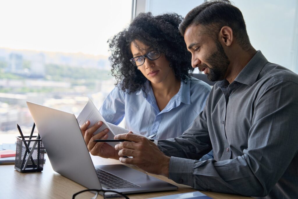 Two people sit at a desk by a window, looking at documents and a laptop, appearing to discuss work.
