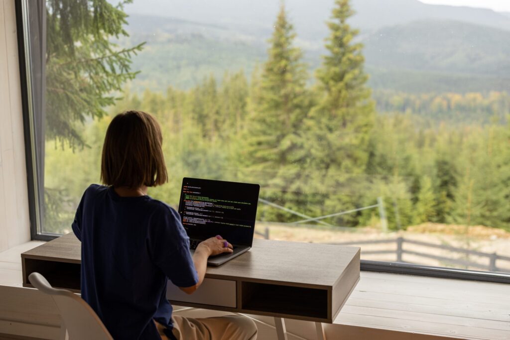 A person sits at a desk by a large window, working on a laptop with code displayed, overlooking a forested, mountainous landscape.