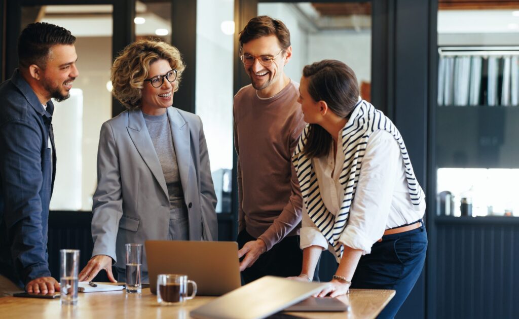 Four people are gathered around a table with a laptop, smiling and discussing marketing strategy services in a modern office setting.
