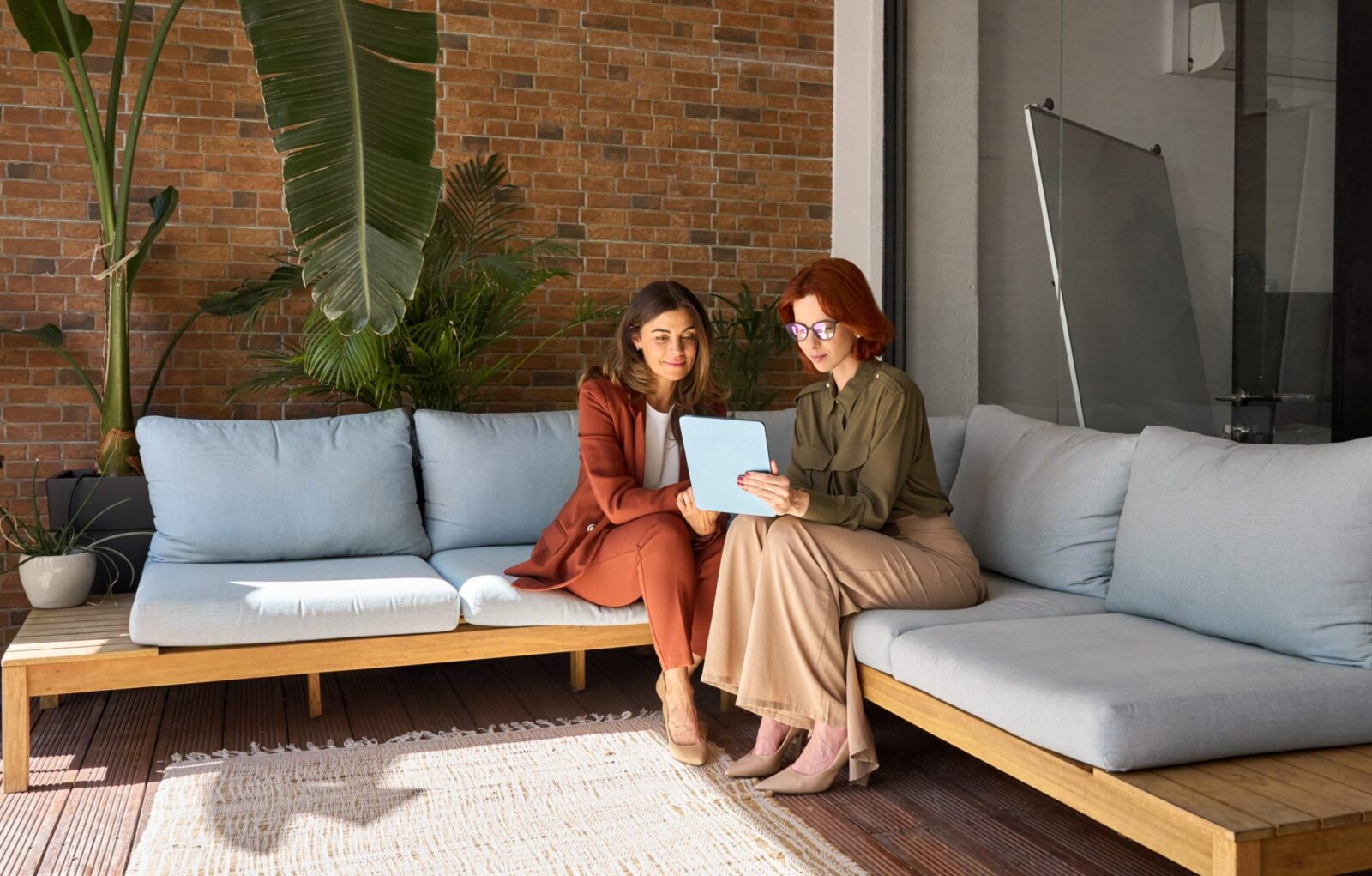 Two women sit on an outdoor sofa, looking at a tablet together. There are plants and a brick wall in the background.