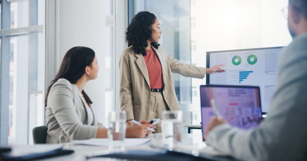 A woman stands and points to graphs on a presentation screen during a business meeting, while two colleagues listen and take notes.