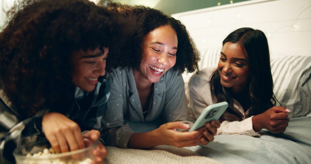 Three young women lie on a bed together, laughing and looking at a smartphone—perhaps brainstorming ideas for their media agency—while one holds a bowl of popcorn.