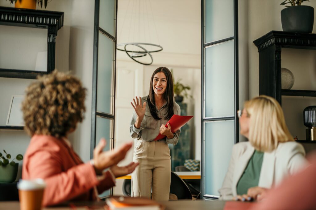 A woman enters a modern office, smiling and waving, while two seated colleagues turn to greet her.