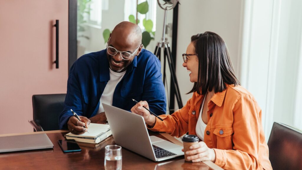 Two people sit at a desk with a laptop, notebook, and drinks, collaborating and smiling in a well-lit office space.