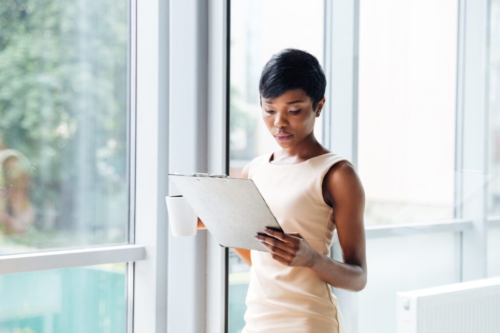 A woman stands by large windows holding a cup in one hand and reading from a clipboard in the other.