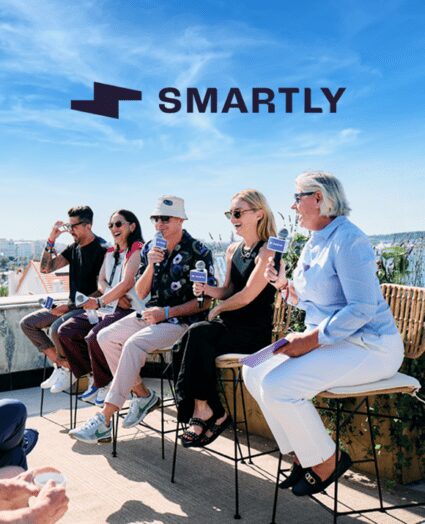 Five people sit on stools outdoors holding microphones under a blue sky, with "SMARTLY" and a logo visible above them.