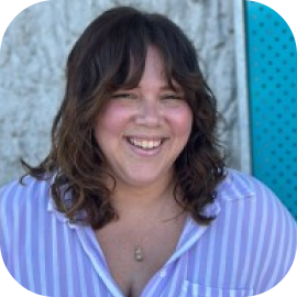 A woman with shoulder-length brown hair and bangs smiles, wearing a light purple striped shirt, standing in front of a textured wall and a blue perforated panel.