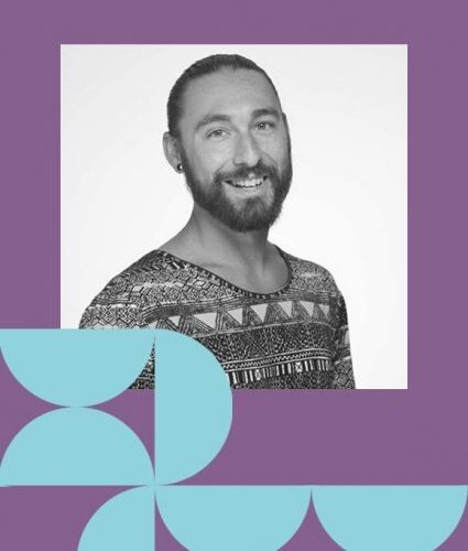 A bearded person with tied-back hair smiles at the camera, wearing a patterned shirt. The photo is framed by geometric shapes on a purple background.