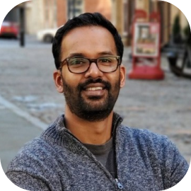 A man with glasses and a beard, wearing a gray zip-up sweater, is smiling while sitting outdoors on a cobblestone street.