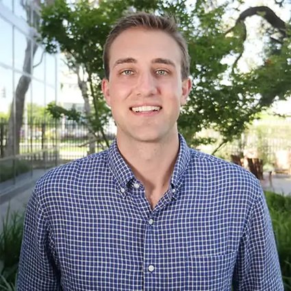 A man in a blue checkered shirt stands outdoors, smiling at the camera with trees and a building in the background.
