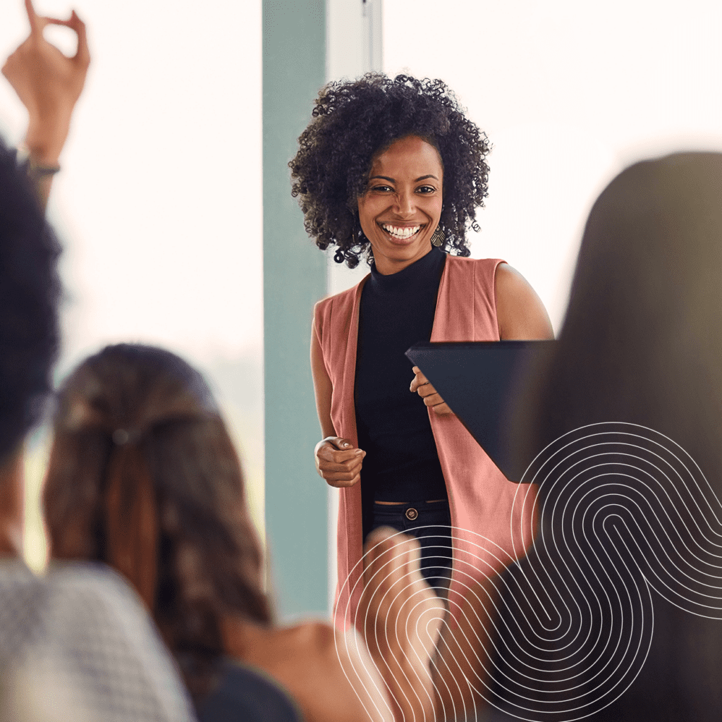 A woman stands at the front of a room holding a folder, smiling at a group of people raising their hands.