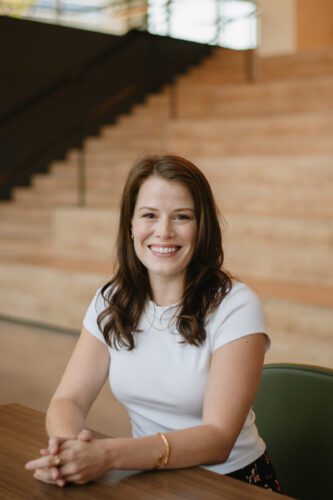 A woman with long brown hair, wearing a white short-sleeve top, sits at a table and smiles, with wooden steps and a railing in the background.