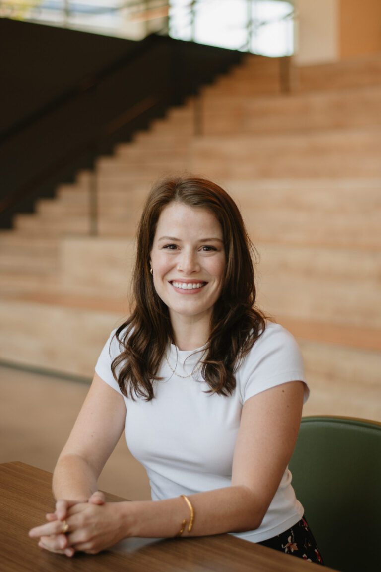A woman with long brown hair, wearing a white short-sleeve top, sits at a table and smiles, with wooden steps and a railing in the background.