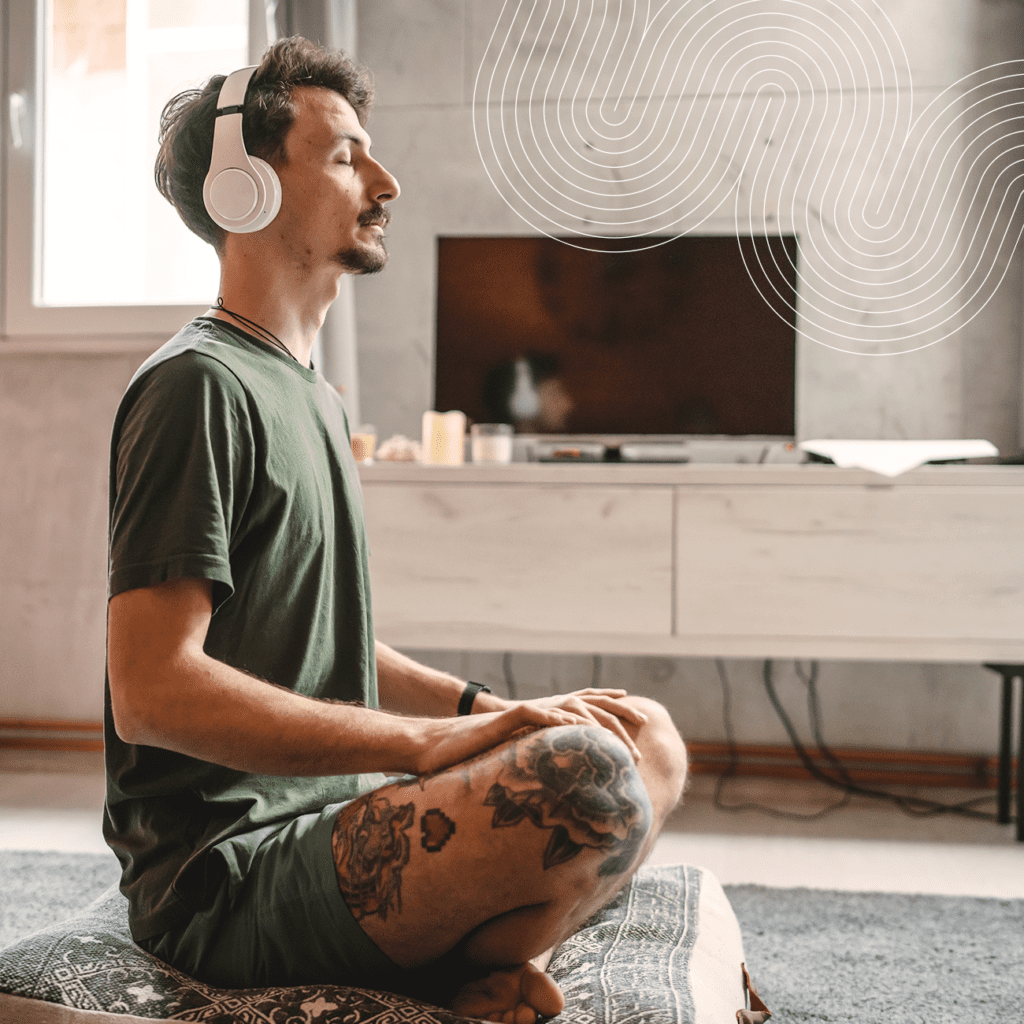 A man with headphones sits cross-legged on a cushion, eyes closed, meditating in a living room with a TV and modern decor.
