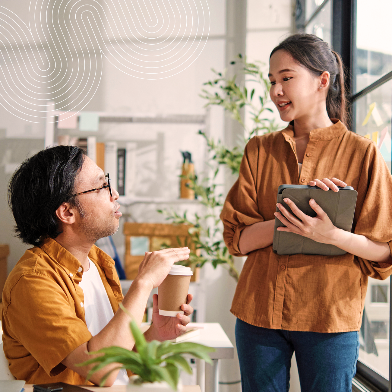 A man holding a coffee cup talks to a woman holding a tablet while standing indoors near large windows and plants.