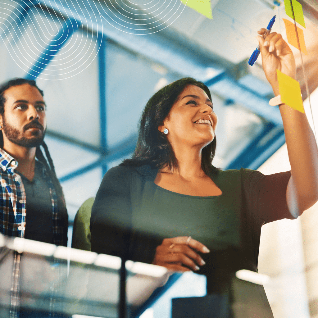 Two people in an office collaborate, with one person writing on sticky notes on a glass wall while the other observes.