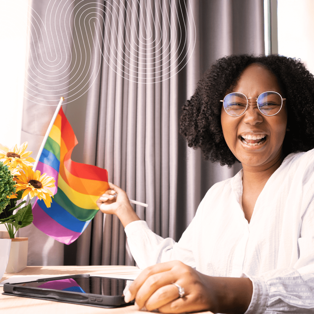 Smiling person wearing glasses holds a rainbow pride flag at a desk with a tablet and a vase of sunflowers, in front of a window with gray curtains.